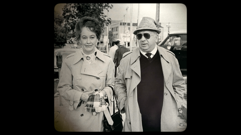 Black and white photo of an older couple in trench coats, walking side by side outdoors.
