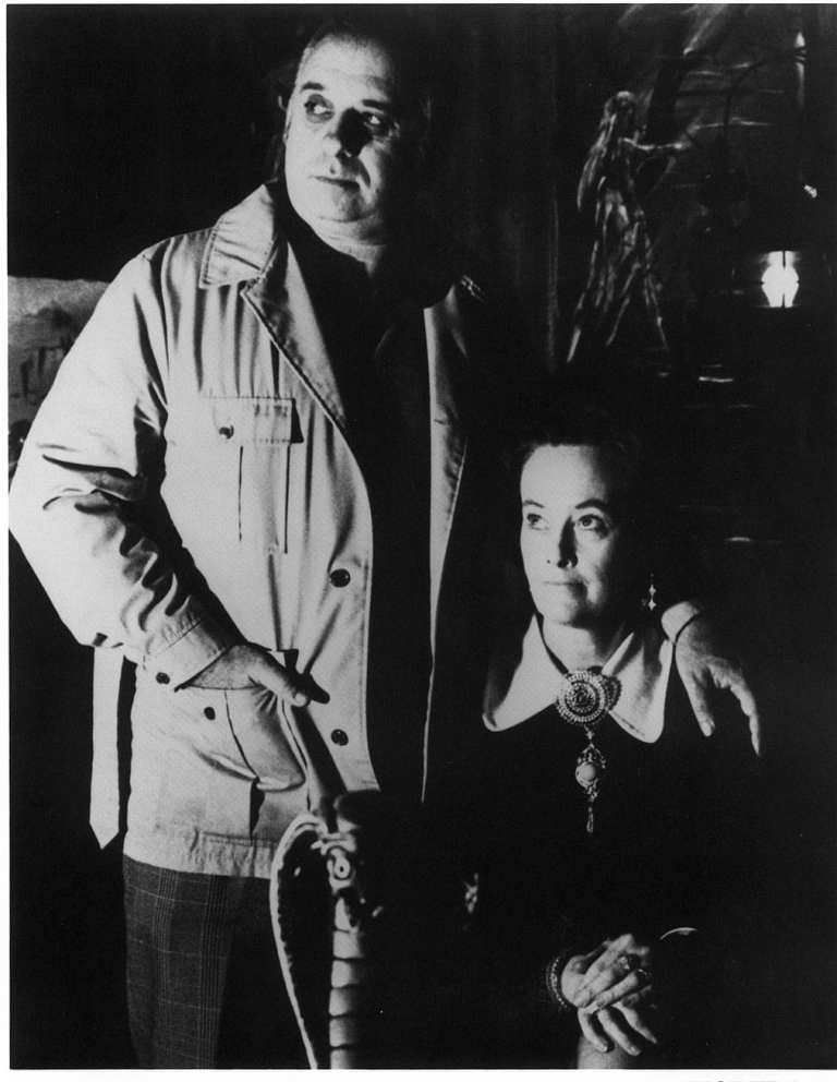 Black and white photo of a man standing and a woman seated indoors.